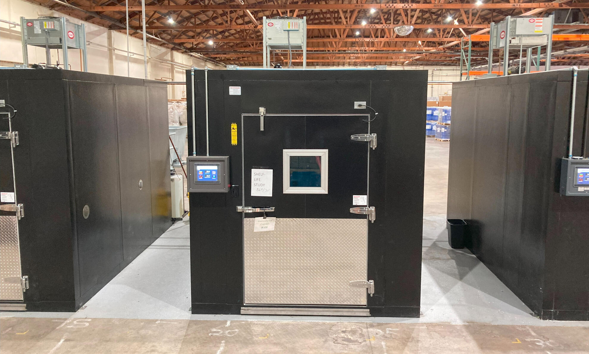 Custom Walk-In Environmental Test Chamber A black walk-in environmental test chamber with a digital control panel and diamond-plated door panel, designed for precise testing in a large industrial facility.